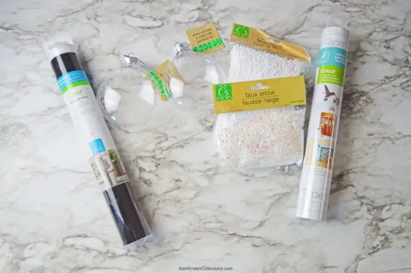 An overhead view of some of the supplies to make DIY Christmas fillable ornaments, such as plastic ornaments, faux snow filler balls, and permanent adhesive vinyl.