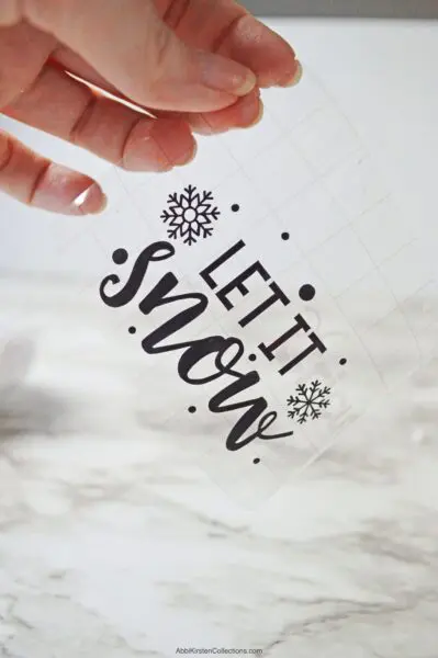 A close-up of Abbi Kirsten’s hand holding clear transfer tape with black vinyl adhered to it. The vinyl says “Let it Snow” and is a free SVG file.