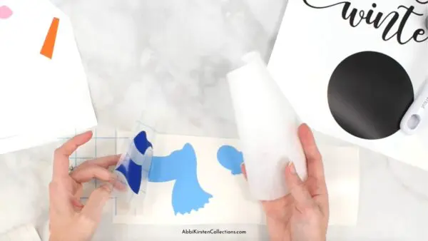 Abbi Kirsten's hands carefully remove transfer tape from a layer of the snowman's scarf. Other vinyl layers lay nearby. 