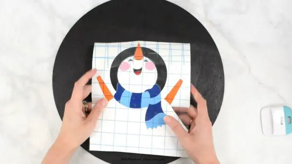 Abbi's hand line up the many layers of the snowman design onto the black circular wood blank. 