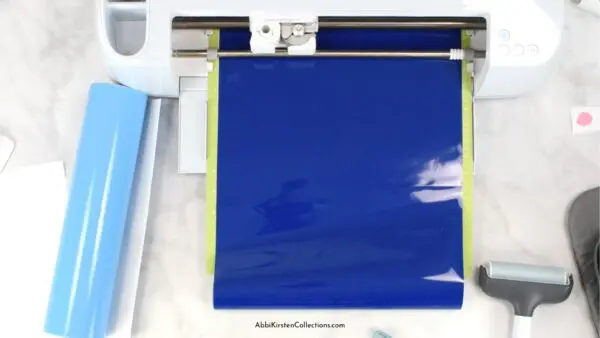 Dark blue vinyl on a mat about to be fed into a Cricut machine for cutting layers of a SVG design. Tools and more vinyl lay around the Cricut machine. 