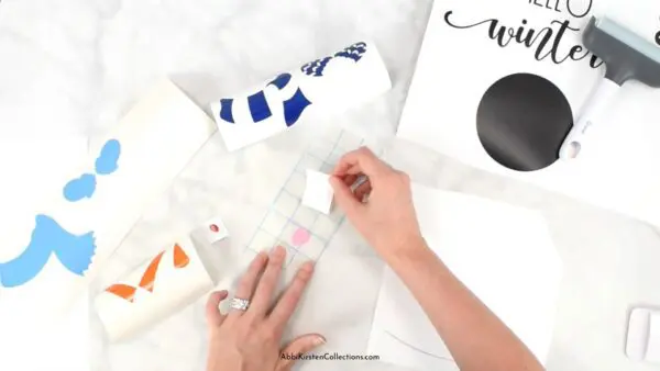 Abbi Kirtsen's hands apply transfer tape to a piece of layered vinyl for her seasonal sign crafts. Around her are other cut layers and printed designs. 