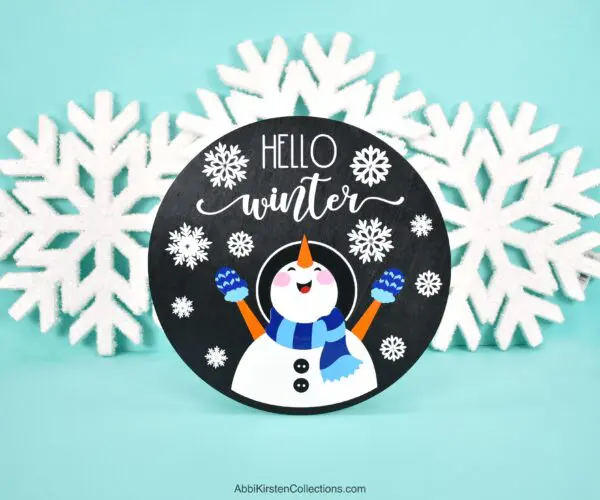 Image shows a black wood sign in front of decorative snowflakes. The sign has a snowman and the phrase "hello winter." The tutorial teaches how to layer vinyl on wood signs with your Cricut machine. 