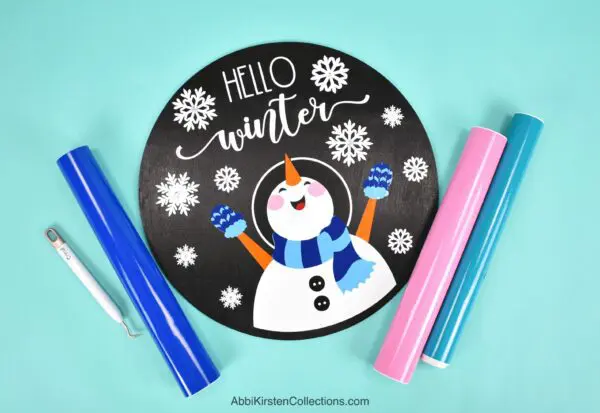 A handmade circular wood sign showing a snowman happy in snowflakes with the words "hello winter" above it. Markers and a weeding tool lay off to the side. 