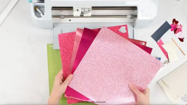 Abbi Kirsten's hands hold glitter cardstock in a fan above a Cricut cutting machine and mat. A Valentine's Day card lays to the right.