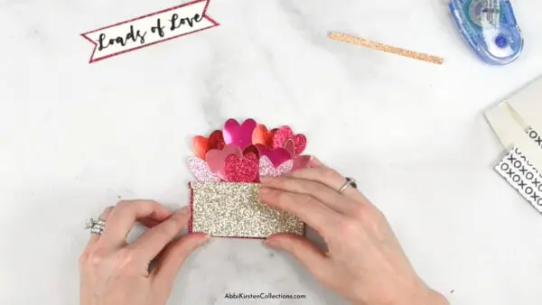 An overhead view of Abbi Kirsten's hands using glitter cardstock heart cutouts and layering them with glue.