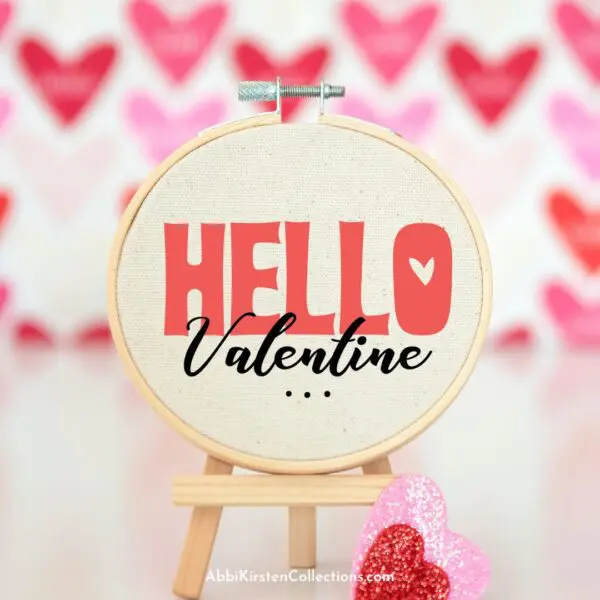 An embroidery hoop holds canvas with the text "Hello Valentine" printed on the front using a Cricut machine and SVG files. Out of focus behind the craft are Valentine's Day heart decorations. 