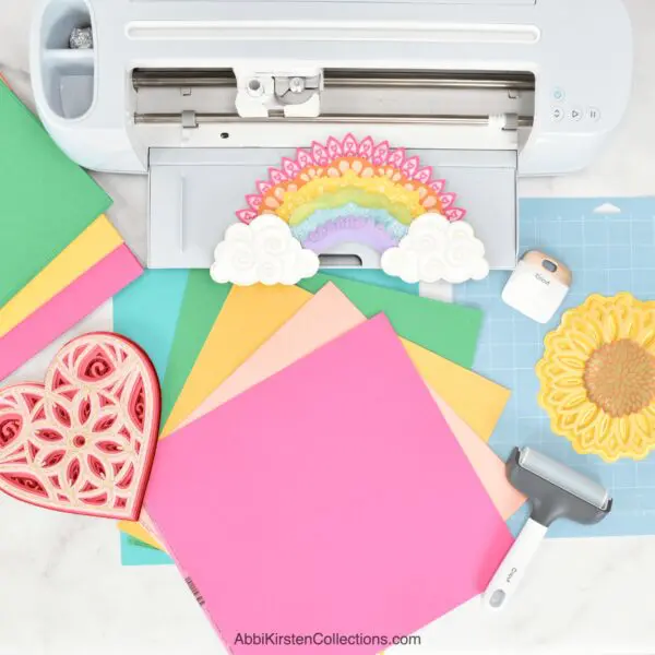The image shows colorful cardstock next to a Cricut machine with several paper craft projects cut out.