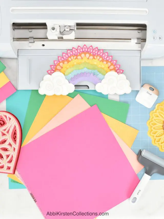 The image shows colorful cardstock next to a Cricut machine with several paper craft projects cut out.