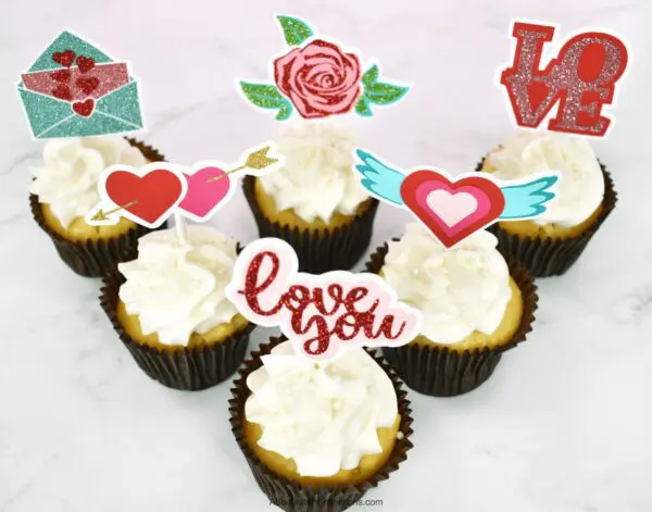 A set of six white cupcakes in a triangle formation each has a different Valentine's Day cupcake topper made with a Cricut machine.  The toppers are two hearts pierced by an arrow, a glittery rose and greenery, an open envelope with hearts, the word “Love,” a heart with wings, and the text “Love you.” 