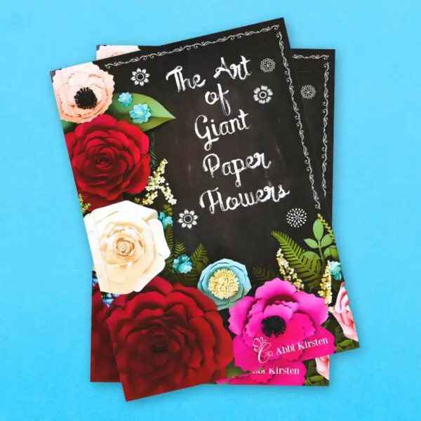 Two copies of a black book with paper flowers on the cover are stacked on a blue background. The title says, "The Art of Giant Paper Flowers."