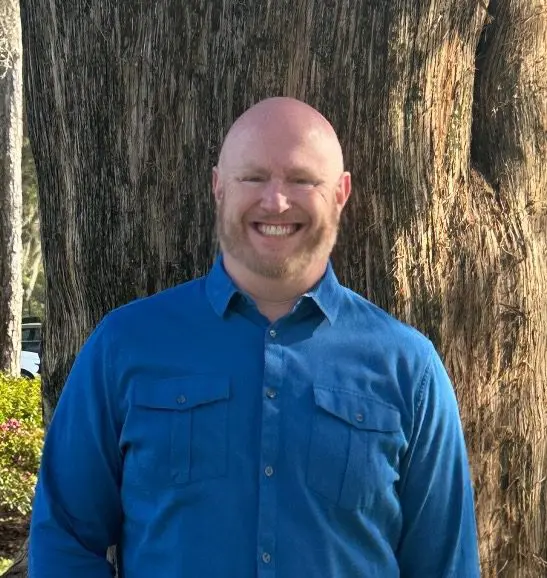 Michael's headshot. He's wearing a blue collard shirt and standing outside, in front of a large tree.