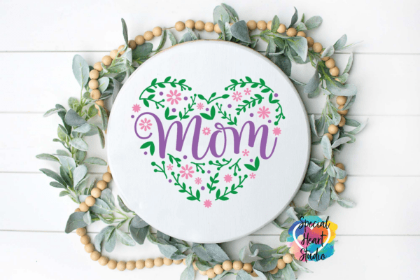 The image shows a floral Mom SVG cut file in pink and purple on a white wood sign. Get this free SVG file here.