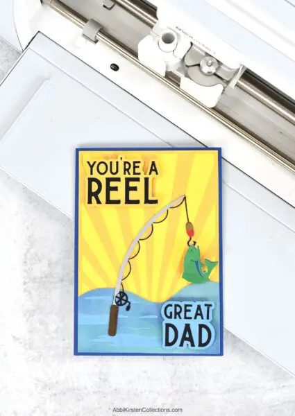 The image shows a DIY Father's Day card on a Cricut machine made from cardstock and iron on vinyl. The card reads, You're A Reel Great Dad. 