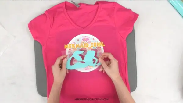 The image shows a pink t-shirt with a third layer of vinyl being placed down to iron-on.