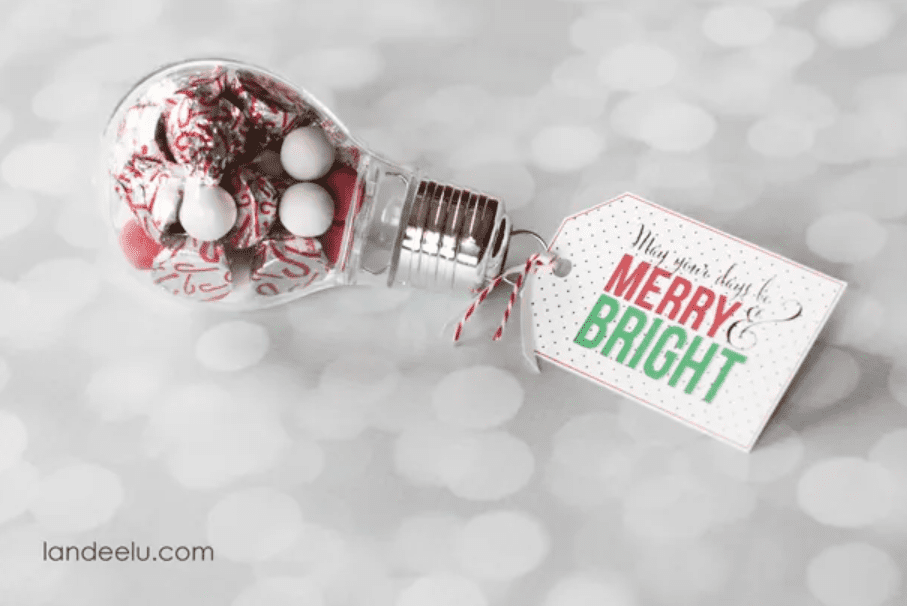 A fake light bulb is filled with Christmas-colored candies. A festive homemade tag on the bulb says, “May your days be merry and bright” in green, red, and gold. The bottom of the picture has the words “Quick and Easy Gift Idea landeelu.com.