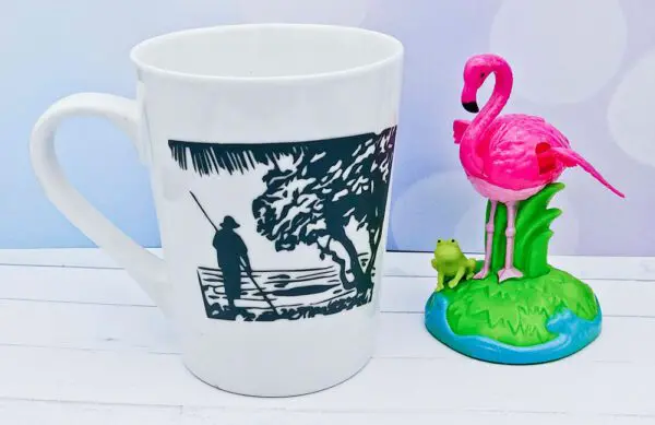 White mug with landscape photo of person on boat and tree with pink flamingo and frog decoration beside mug.