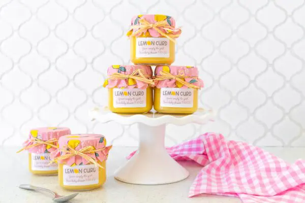 Lemon curd jars with custom labels and covers on white cake stand with pink and white checkered dish towel.