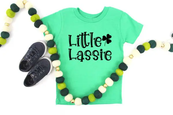 Green t-shirt with shamrock and text Little Lassie, child's black sneakers, and green and white pom pom garland, offered as a free SVG.