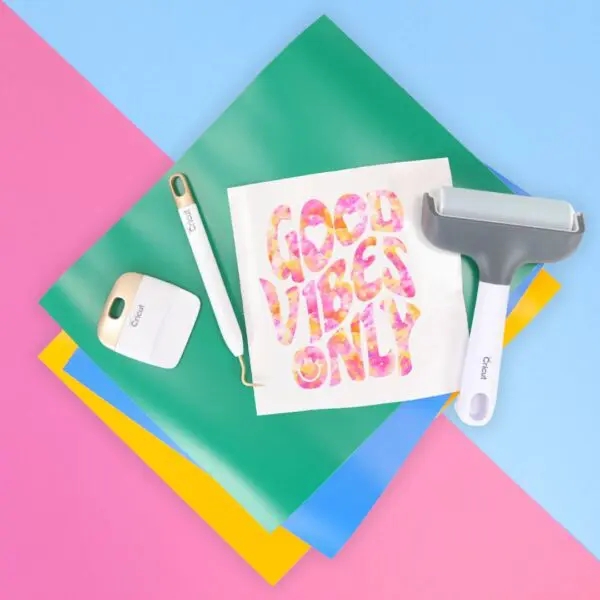 Cricut crafting materials, vinyl, brayer tool and a design that reads Good Vibes Only laying on a pink and blue table.