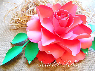 A pink scarlet paper rose, resting on green paper leaves. Notice the unique cupping of the petals. You can make these delicate flowers during your crafting time. 