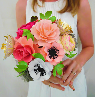 A close-up of a bridal and wedding bouquet full of multi-colored paper flowers and green leaves and vines made using a cutting machine, held by Abbi Kirsten. 