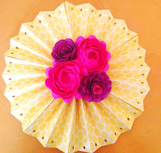 On a wooden tabletop lays a rosette paper fan created with yellow and white scrapbooking paper. The center has been decorated with pink and purple paper peony and dalhia flowers. 