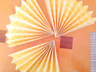 Three side of a four-sided fan lay on a wooden table, ready to be made into a rosette paper fan. 