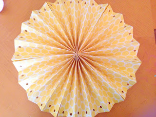 A round rosette fan made of yellow and white scrapbooking paper. Each edge has a hole punched into it. 