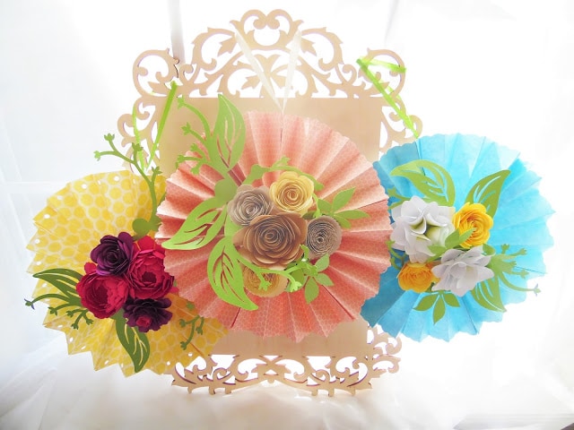 The images shows DIY paper fans made into rosette shapes with paper flowers. Paper flowers adorn the fans in the center, and a lace cut-out acts as backdrop.  