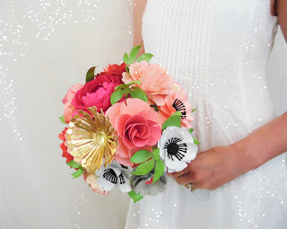 A close-up of a bride holding a paper flower bouquet at waist height. The flowers are pink, gold and white, and have paper greenery as well. 
