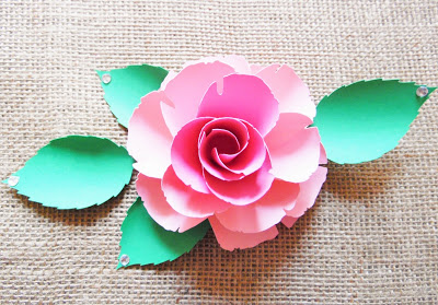 A bird's eye view of an open pink Ruby Rose paper flower and paper leaves. The flower sits on a tan canvas. 