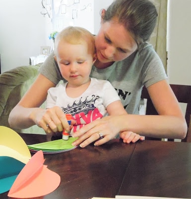 Abbi and her daughter - a young blonde toddler sitting in her lap - apply a glue stick to a folded piece of green paper.