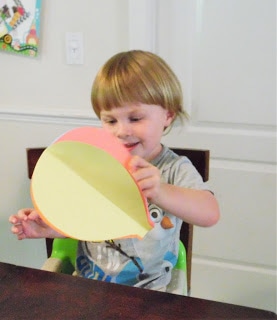 A young boy with shaggy blonde hair sits in a chair, holding an almost-complete paper hot air balloon craft, made from orange and yellow paper.