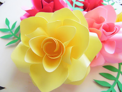 Bright yellow and pink paper made the Ansley roses blooming on a white table. Light green paper foliage peeks out from around the three springtime paper flowers. 