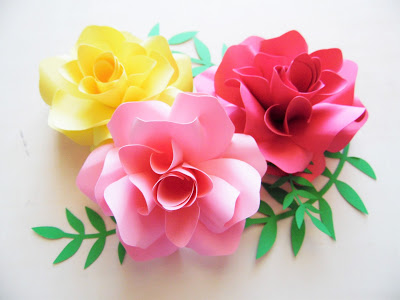 Three paper Ansley roses in deep pink, light pink, and yellow are bunched together on an empty table with paper fern-like foliage connecting them all. 