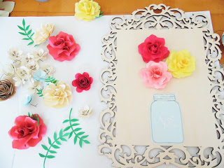 Using a wooden frame as a base, pre-made paper flowers and leaves are glued on to the frame, above the cutout of a paper mason jar. 