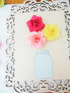 Three paper flowers are glued above a paper mason jar on a wooden decorative frame. 