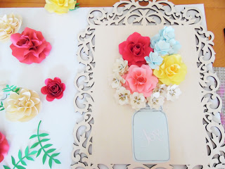 Smaller, pre-made paper flowers are ready to be glued above a blue paper mason jar cut-out on an unfinished decorative wooden frame. 