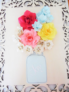 Various papercraft flowers are arranged and glued above a paper mason jar on an unfinished wood frame. 