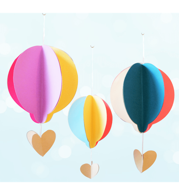 Three colorful paper hot air balloon crafts hang from the ceiling with string. The hot air balloon crafts are made with pink, yellow, blue, and white paper, and have paper hearts hanging from the base.