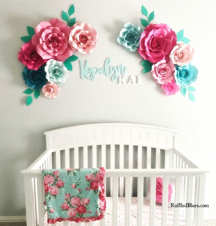 Colorful paper flowers in shades of pink, mint, and blue hang over a baby girl's crib on the white wall of her nursery.