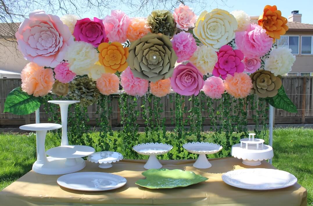 A variety of colorful giant paper flowers make up this background, set up at an outdoor party. Vines of grass hang from the paper flowers, creating a perfect photo background. Cake stands and platters are laid out on the table in front of the giant paper flower backdrop.