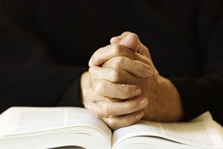 Folded hands rest on an open Bible, praying.