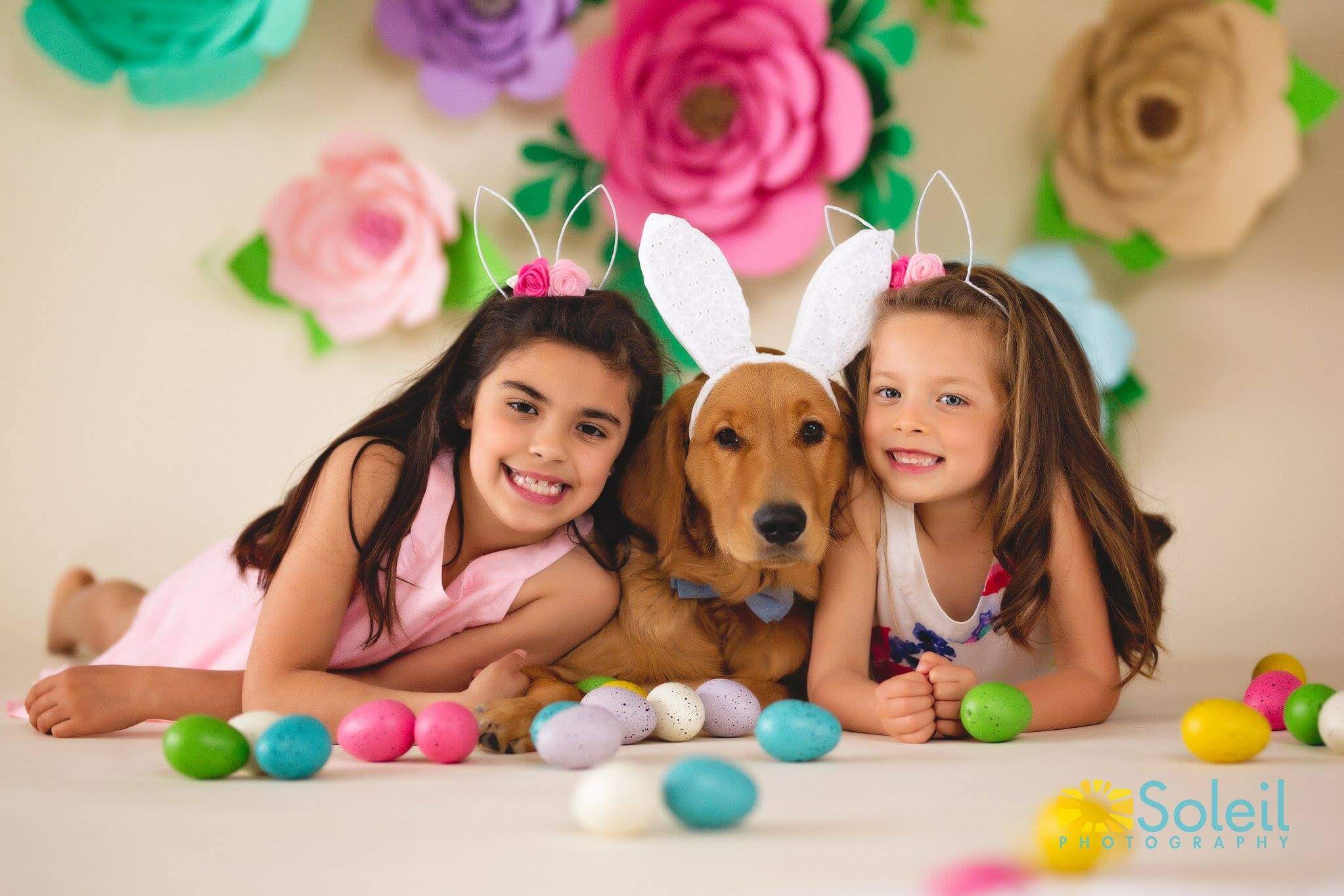 Two young girls wearing Easter bunny ears post with their pet dog, also wearing a pair of Easter bunny ears. In the background, giant paper flowers hang on a white wall. Easter eggs are scattered on the floor around the girls and dog.