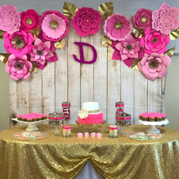 Giant paper flowers in various shades of pink adorn a wooden wall behind a party dessert table. The table is covered with a glittery gold tablecloth under cakes and cupcakes.