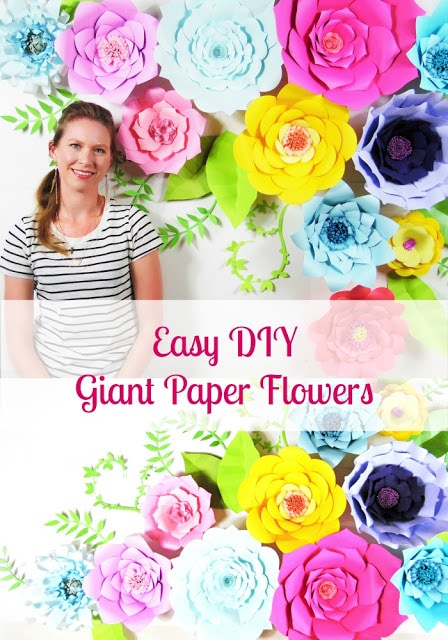 A woman stands in front of a colorful paper flower backdrop made with giant paper flowers in purple, yellow, pink, and blue. A banner across the image says "Easy DIY Giant Paper Flowers"