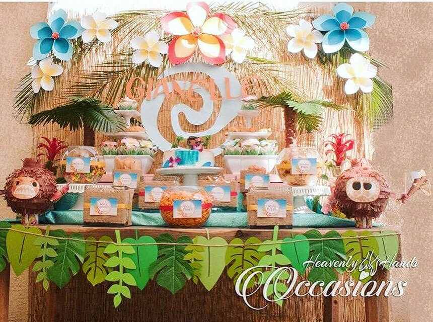This Moana-themed snack table includes tropical paper flowers hanging on the wall in shades of white, pink, and blue. A tropical leaf banner hangs on the front of the table, which is filled with boxes of snacks.
