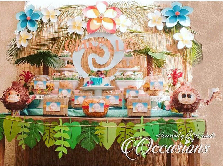 This Moana-themed snack table includes tropical paper flowers in shades of white, pink, and blue hanging on the wall. A tropical leaf banner hangs on the front of the table, which is filled with boxes of snacks.