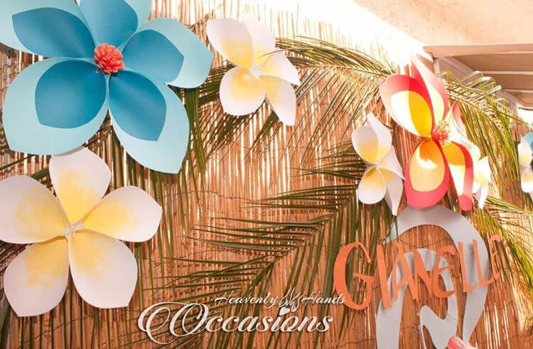 A close up look at the Hawaiian flowers mounted on a seagrass background at a Moana-themed birthday party. The paper flowers are made with blue, white, yellow, and coral colored paper.